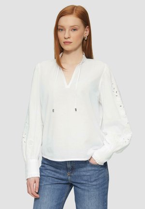Witte blouse met lange mouwen, V-hals, trekkingdetail en geborduurde bloemenaccenten op de mouwen. Gecombineerd met blauwe jeans.