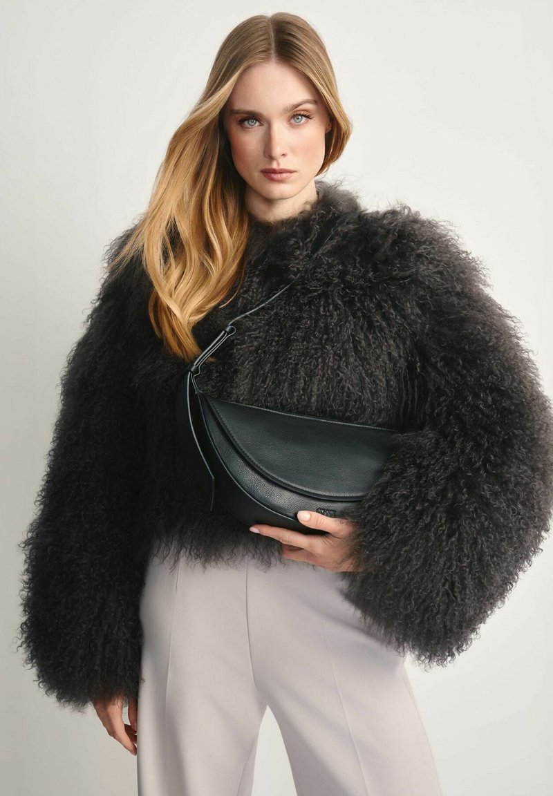 Kobieta z długimi blond włosami, ubrana w puszystą czarną kurtkę i jasne spodnie, trzyma czarną skórzaną torebkę na ramię.