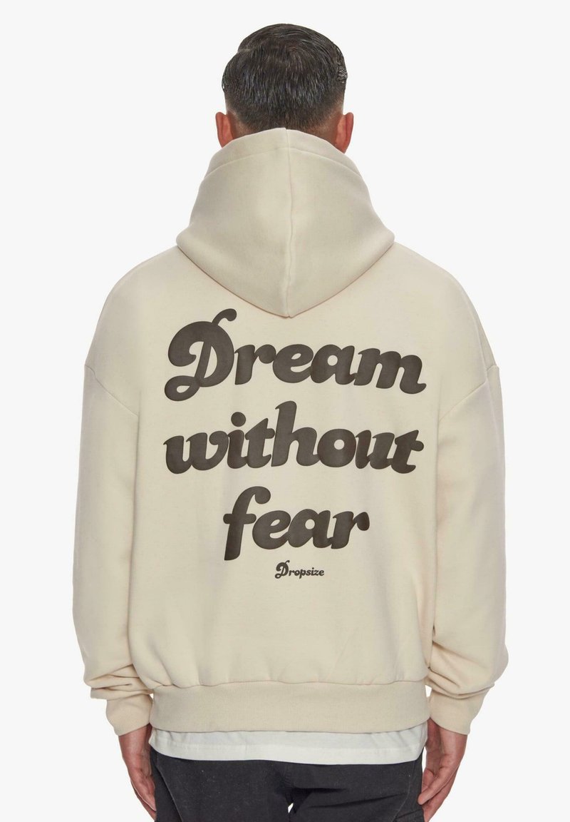 Cremfarbener Kapuzenpullover mit Kängurutasche, auf der Rückseite mit "Träume ohne Angst" in kräftigen dunkelbraunen Buchstaben bedruckt.