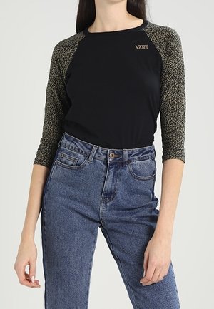 Mujer con una camiseta negra de Vans con estampado de leopardo y mangas 3/4, y jeans azules de talle alto, de pie frente a un fondo liso.