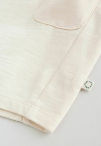 Nahaufnahme eines Ärmels aus beigem Stoff mit einem kleinen weißen Etikett, das grüne kreisförmige Schrift auf einer weißen Oberfläche zeigt.