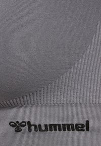 Zbliżenie na szary materiał w prążki z czarnym logo hummel i symbolem pszczoły wyhaftowanym w dolnej części.