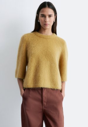 Kvinde med langt mørkt hår iført store øreringe, en blød sennepsgul sweater med trekvartærmer og højtaljede brune bukser, hænderne i lommerne.