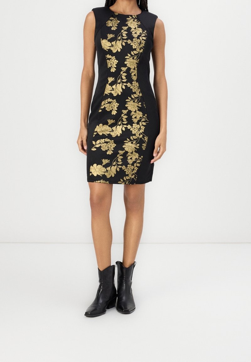 Femme portant une robe noire sans manches avec des motifs floraux dorés sur le devant, associée à des bottines noires.