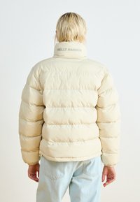 Puffer jacket in light beige with horizontal quilted design, high collar, and "HELLE HANSEN" logo at the neck. Paired with light blue jeans.