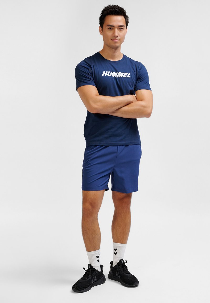 Marinblå joggingtröja med vitt "HUMMEL"-logotyp, parat med matchande marinblå shorts. Modellen bär svarta sneakers och vita strumpor med svarta detaljer.