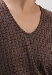 Tejido marrón texturizado con un patrón de cuadrícula, que presenta un diseño de cuello en V. El material parece suave y ligeramente elástico.