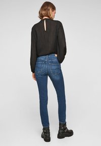 Blusa negra de manga larga con textura transparente, con escote en forma de llave en la parte posterior, combinada con jeans ajustados azules que cuentan con dos bolsillos traseros y un acabado suave.