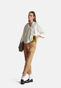 Blusa bianca oversized con scollo a V, abbinata a pantaloni beige affusolati. Inclusi mocassini neri, orli arrotolati e una borsa verde con texture.