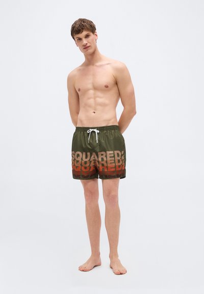 Νέος άνδρας όρθιος, ξυπόλητος, φορώντας μαγιό ελιάς με διαβαθμισμένο κείμενο "SQUARED" και λευκό κορδόνι σε απλό φόντο.