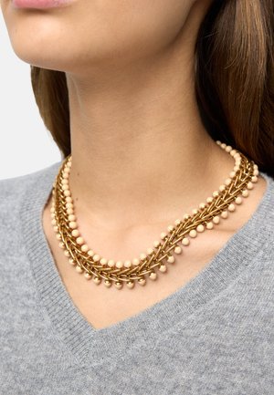 Zbliżenie na osobę noszącą warstwowy naszyjnik z łańcuszków ze złota z beżowymi koralikami na jasnoszarym swetrze z dekoltem w serek.