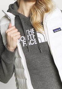 Gilet imbottito bianco a trapunta con chiusura zip, indossato sopra un felpa grigia con grande testo bianco. Il gilet presenta un'etichetta con il marchio sul petto.