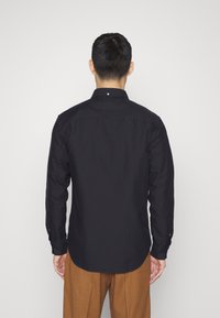 Camicia nera a maniche lunghe con colletto classico, chiusura a bottoni e una consistenza liscia, abbinata a pantaloni color ocra.