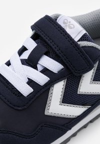 Navy sneaker with mesh and suede materials. Features white laces, Velcro strap, and a zigzag design on the sides. Textured gray lining.