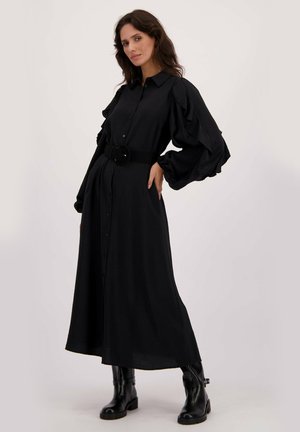 Vrouw in een lange zwarte jurk met knopen, pofmouwen en een brede riem, gecombineerd met zwarte enkellaarsjes, staand tegen een vlakke achtergrond.