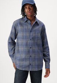 Blå rutete skjorte med teksturert stoff, knappestolpe foran og lange ermer. Brukt med mørk denimbukser og en blå caps.