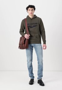 Oliwkowy hoodie z czarnym logo, niebieskie dżinsy i czarne buty. Brązowa torba na ramię z dwiema przednimi kieszeniami, gładka skóra.