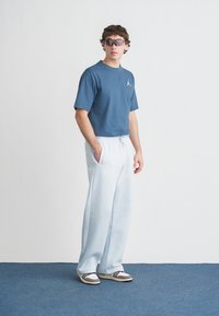 Φαρδιά γαλάζια μπλούζα με λογότυπο, συνδυασμένη με ανοιχτό μπλε φόρμα και αθλητικά παπούτσια με γκρι λεπτομέρειες. Το μοντέλο φορά γυαλιά ηλίου και στέκεται όρθιο.