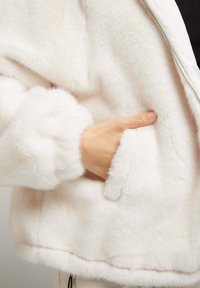 Veste en fausse fourrure blanche avec une texture douce, dotée d'une fermeture éclair et d'une poche latérale, conçue avec des poignets et un ourlet court.