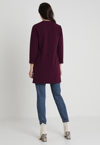 Mörkröd långärmad tunika med rak skärning och uppvikta ärmar. Matcher med blå jeans och vita ankelstövlar med ormskinnsmönster.