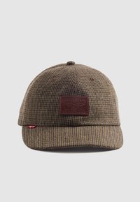 Brun hounds-tooth keps med en böjd skärm och ett texturerat tyg. Har en brun läderlapp med varumärkeslogotyp på framsidan. Röd etikett på sidan.