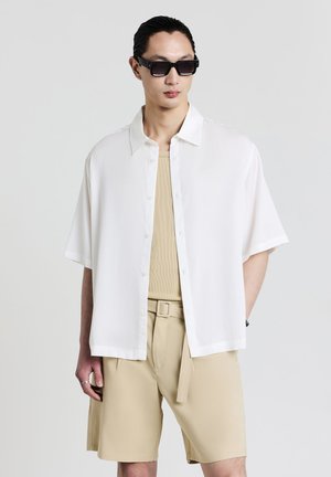 Man wearing black sunglasses, loose white short-sleeve button-up shirt, beige tank top, and beige belted shorts against a plain background.