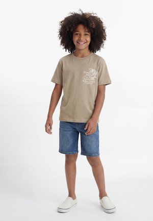 Enfant souriant aux cheveux bouclés portant un t-shirt beige imprimé, un short en jean et des chaussures blanches à enfiler, debout devant un fond blanc uni.