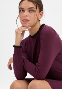 Sukienka z długim rękawem w głębokim fioletowym materiale z czarną falbaną przy dekolcie i mankietach. Charakteryzuje się prążkowaną teksturą i dopasowanym krojem.