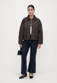 Woman wearing a dark brown quilted jacket, white top, dark blue jeans, black flats, gold hoop earrings, holding a small black handbag.