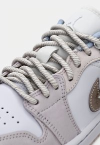 Gros plan d'une basket blanche et rose clair avec des lacets tressés beiges épais noués à travers des œillets métalliques sur une tige en cuir souple.