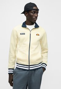 Veste à fermeture éclair jaune clair avec col marine, poignets rayés et logo brodé ; présente une texture lisse et un design sportif.