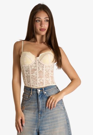 Donna con lunghi capelli castani che indossa un top corsetto in pizzo crema e jeans blu sbiaditi a vita alta con dettagli usurati.