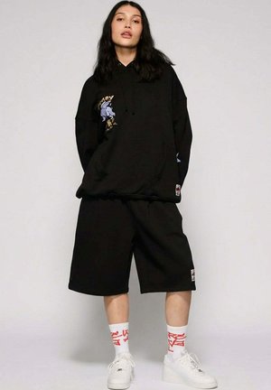 Młoda kobieta o długich ciemnych włosach, ubrana w oversize'ową czarną bluzę z kapturem i szorty, białe skarpetki z czerwonym wzorem oraz białe sportowe buty, stoi.