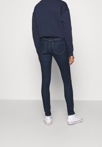 Person wearing dark blue skinny jeans, navy sweatshirt, and white high-top Converse sneakers standing against plain background.