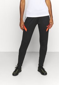 Zwarte sportbroek met oranje accenten, tapered fit, elastische tailleband, zijkanten met strepen, gecombineerd met zwarte sportschoenen met groene details.