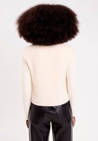 Personne avec de grands cheveux bouclés portant un haut à manches longues crème et un pantalon en cuir noir, vu de dos contre un fond uni.
