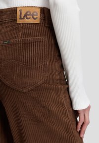 Gros plan sur un pantalon en velours côtelé marron avec des étiquettes de la marque Lee sur la poche arrière, porté avec un haut blanc à manches longues côtelées montrant une main sur le côté.