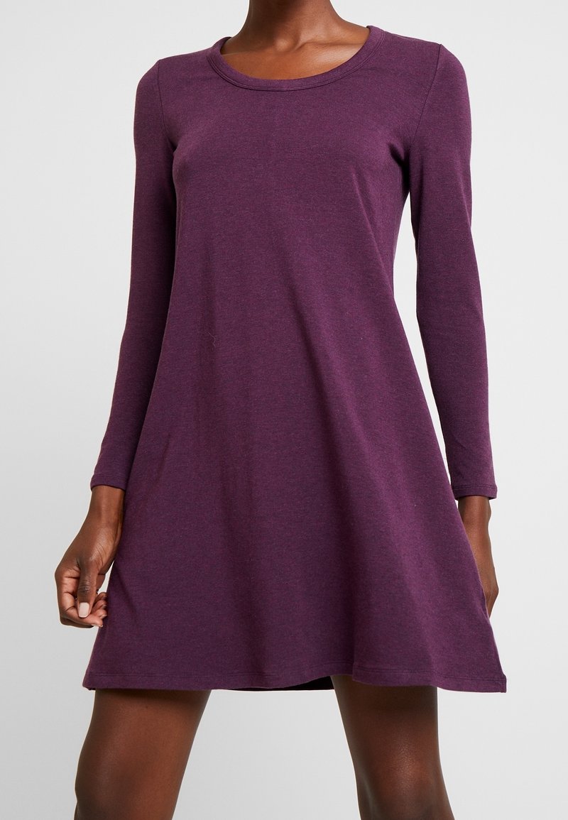 Femme portant une robe patineuse à manches longues, longueur genou, de couleur violet foncé, sur un fond gris clair uni.