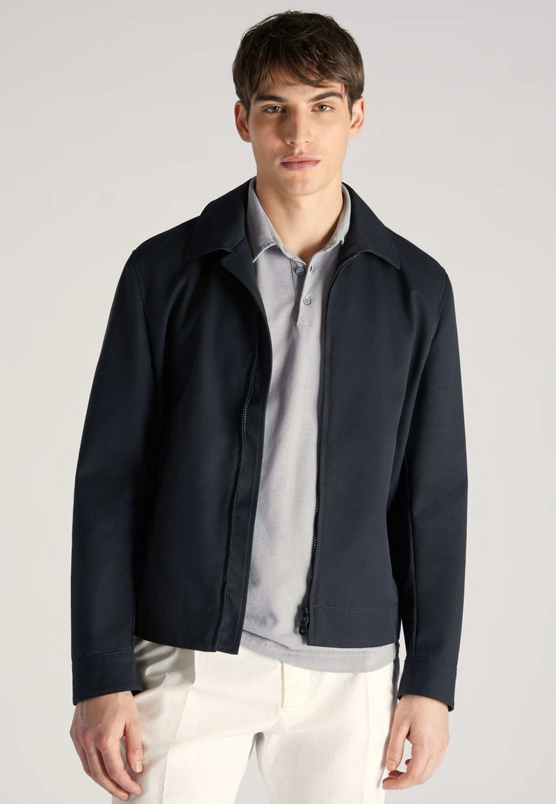 Granatowa kurtka o gładkiej fakturze, z ostrym kołnierzem i zamkiem z przodu. Nosi się ją na szarej koszulce polo, zestawiona z jasnymi spodniami.
