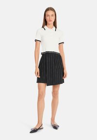 Weißes, geripptes Polohemd mit schwarzen Akzenten, kombiniert mit einem schwarzen, gestreiften Rock mit einer Seitentasche. Schwarze Ballerinas vervollständigen den Look.