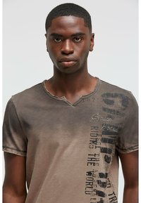 Brown cotton T-shirt with a V-neck, faded texture, and printed text along the side. Features distressed edges and a casual fit.