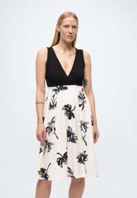 Juoda ir kreminė suknelė su gilia V formos iškirpte, gėlėtu juodu raštu ant šviesaus fono, A formos sijono stiliaus ir lygaus audinio tekstūra.