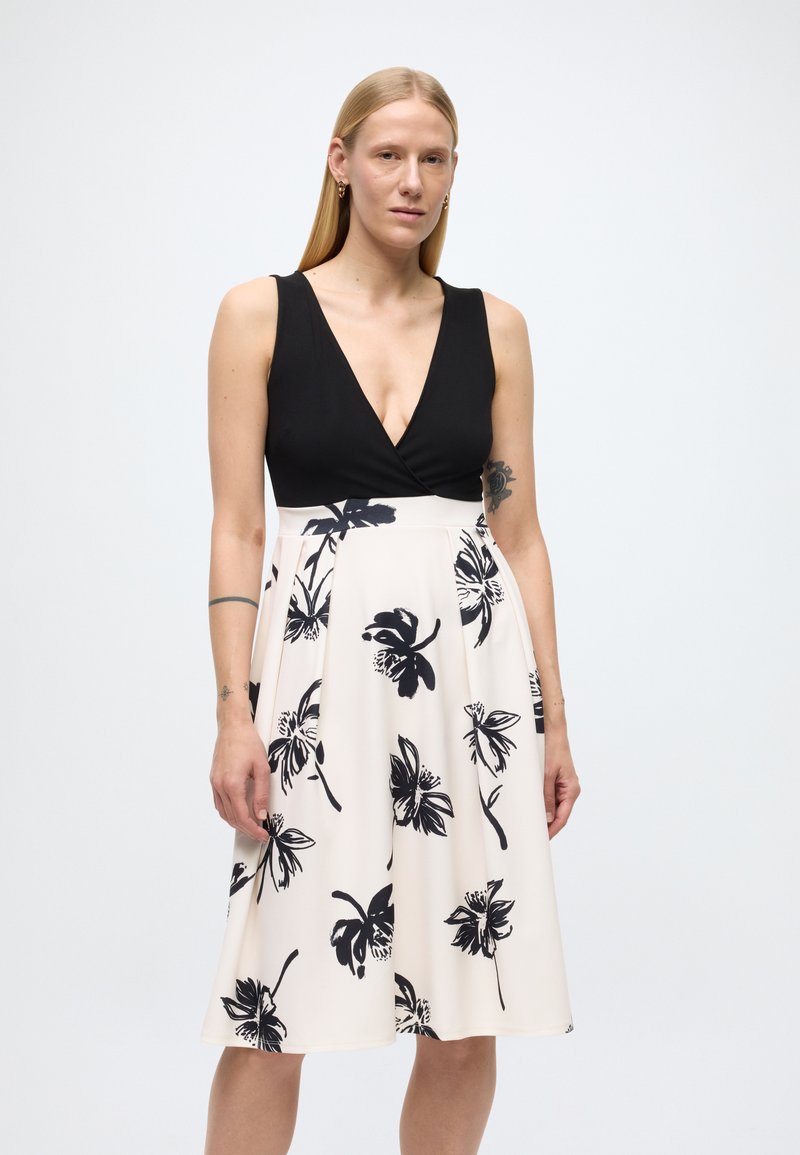 Juoda ir kreminė suknelė su gilia V formos iškirpte, gėlėtu juodu raštu ant šviesaus fono, A formos sijono stiliaus ir lygaus audinio tekstūra.