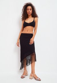 Schwarz gerippter Bralette, kombiniert mit einem kunstvoll gewebten schwarzen Rock, der am Saum mit Fransen verziert ist. Leichte Blockabsatzsandalen runden den Look ab.