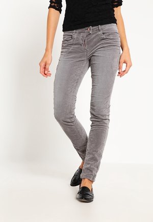 Femme portant un pantalon en velours côtelé gris ajusté, un haut noir avec des manches en dentelle, et des mocassins noirs, debout avec un pied légèrement en avant.