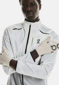 Veste légère blanche avec un col montant, fermeture éclair noire et coutures contrastées. Gants beiges avec un logo et ajustement confortable au niveau des poignets.