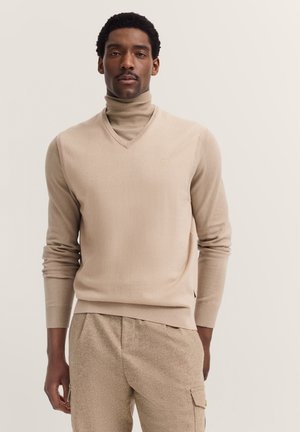 Beige Rollkragenpullover mit V-Ausschnitt, gerippten Bündchen und seitlichen Schlitzen. Die glatte Textur und der enganliegende Schnitt ergänzen die lässig geschnittenen beigen Hosen.
