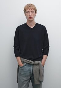 Maglione blu scuro a V con maniche lunghe, abbinato a jeans sbiaditi blu chiaro. Un maglione grigio è annodato intorno alla vita. Sfondo semplice.