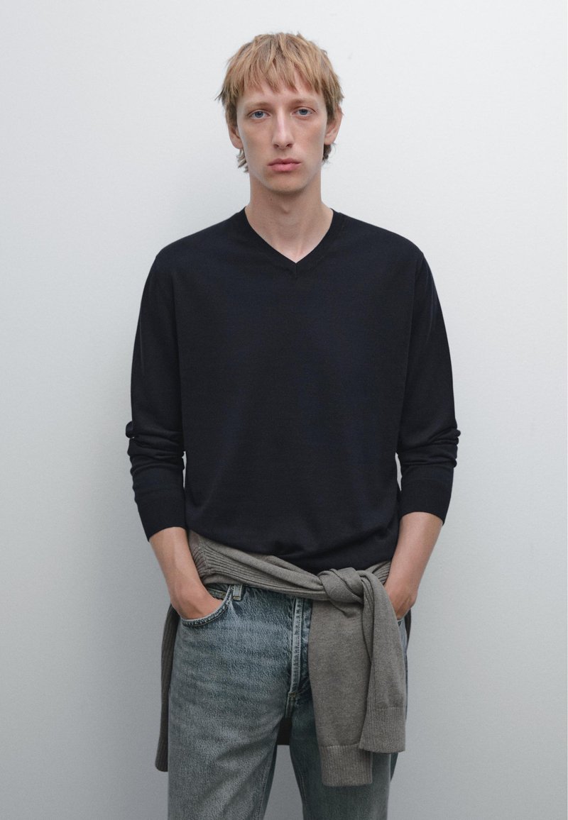 Maglione blu scuro a V con maniche lunghe, abbinato a jeans sbiaditi blu chiaro. Un maglione grigio è annodato intorno alla vita. Sfondo semplice.