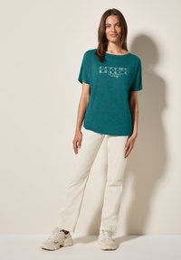 T-shirt teal à manches courtes avec un col rond et un texte graphique blanc. Associé à un pantalon droit écru et des baskets beiges.